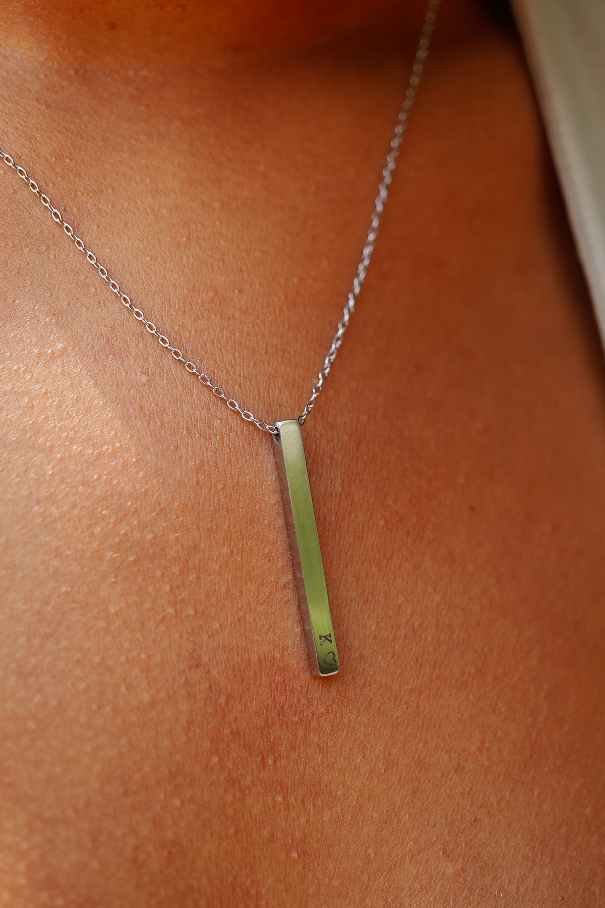 A close-up of a silver bar necklace worn on a person's neck, featuring a polished vertical bar pendant on a delicate link chain.