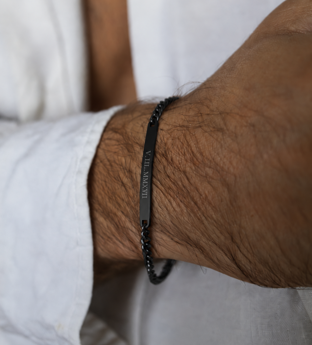 A close-up of a person's wrist wearing a black beaded bracelet with a central black metal plate engraved with roman numerals. The person is wearing a white linen shirt.