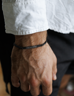 A close-up of a man's hand and wrist wearing a black metal chain bracelet engraved with roman numerals. He is wearing a white collared shirt and black shorts.